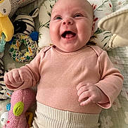 Ava is registered to the contest to win money with this photo: baby, infant, smiling, happy, lying_down, plush_toy, pink_clothing, cream_pants, face, child, cute, joyful, indoors, toy, soft_texture, blanket, patterned, closeup, human, playtime