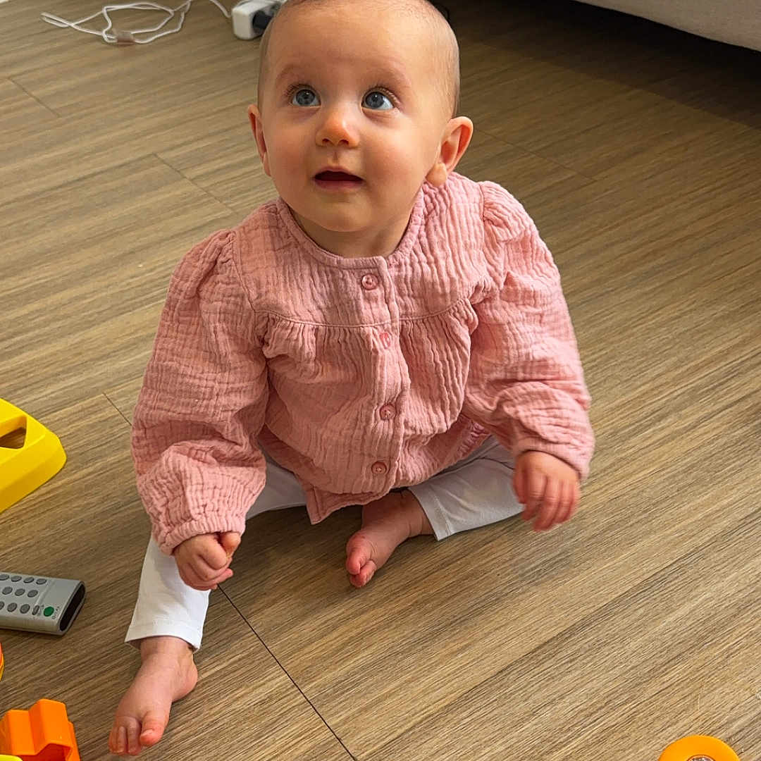 Mencía a rejoint le concours — aidez-le/la à gagner de superbes lots ! baby, blanket, child, colorful_toy, couch, curious, cute, floor, foot, hand, indoor, infant, looking_up, person, pink_shirt, remote_control, sitting, toy, white_pants, wooden_floor