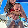 Colton is registered to the contest to win money with this photo: baby, child, woman, beach, umbrella, sunny, smiling, happy, holding, shorts, glasses, outdoor, sand, blue_sky, vacation, family, summer, people, building, casual