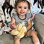 toddler, child, smiling, plush_toy, pink_bow, striped_shirt, shorts, socks, couch, blanket, horse_pattern, indoors, happy, person, portrait, cute, young_child, seated, home, comfort