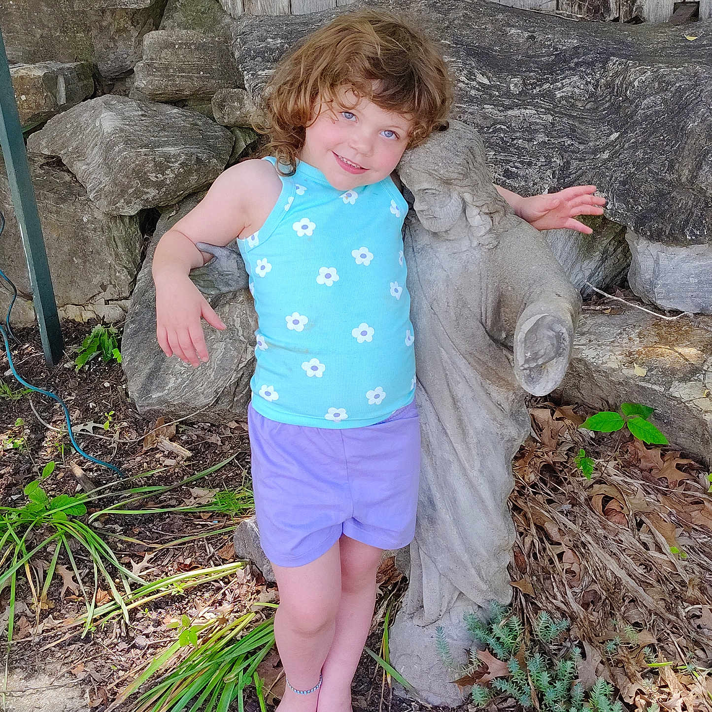 Aryanna joined the competition — help win amazing prizes! barefoot, blue_eyes, casual_clothing, child, curly_hair, garden, greenery, nature, outdoor, plants, portrait, purple_shorts, rocks, smiling, statue, stone, sunlight, tank_top, wooden_fence, young_child