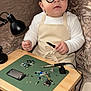 baby, child, goggles, apron, table, electronics, circuit_board, soldering_iron, lamp, couch, indoor, curious, tinkering, technology, tools, small_table, seated, person, closeup, cozy