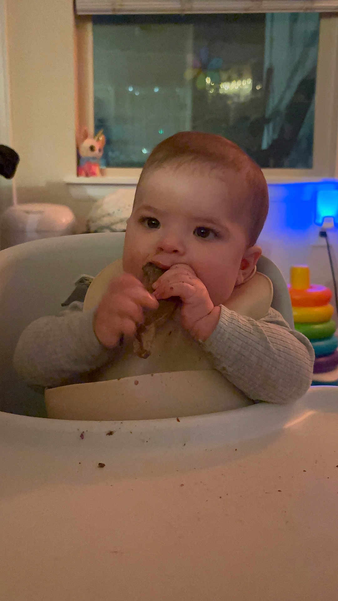 Jasper is registered to the contest to win money with this photo: baby, child, high_chair, food, eating, indoor, toy, window, bib, face, hand, table, sitting, clothing, furniture, head, person, room, home, soft_lighting