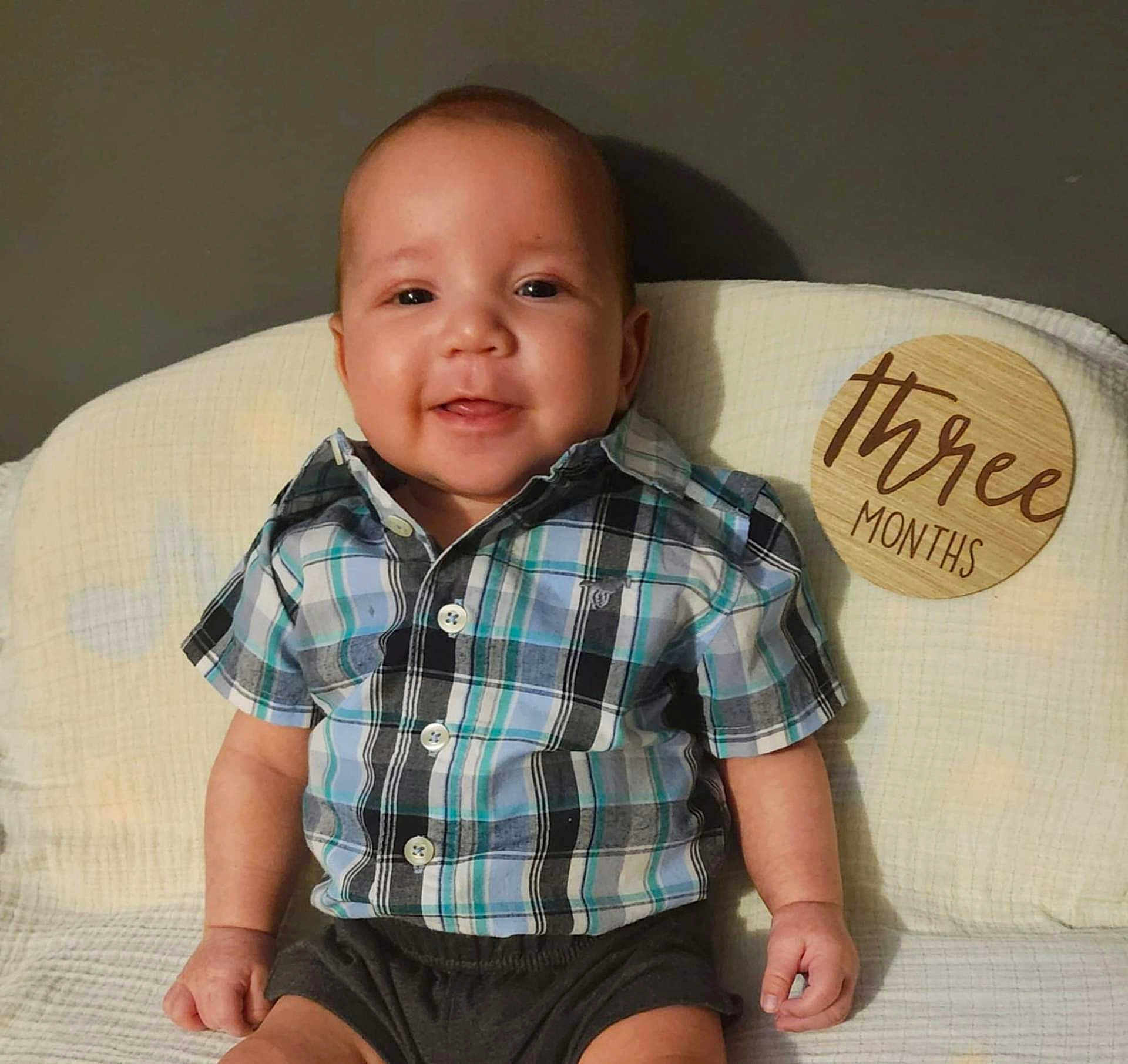 Mason is registered to the contest to win money with this photo: baby, infant, child, smiling, plaid_shirt, shorts, sitting, cushion, white_blanket, wooden_sign, milestone, portrait, indoors, cute, happy, face, head, person, young_child, three_months