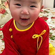 Luana is registered to the contest to win money with this photo: baby, child, smile, red_outfit, yellow_trim, sitting, hands, floral_bedspread, closeup, portrait, indoor, door, door_knob, rosy_cheeks, short_hair, cute, eyes, bedroom, fabric, happy