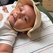 Azael is registered to the contest to win money with this photo: baby, infant, hat, face, smile, lying_down, blanket, striped_blanket, arm, indoor, furniture, drawer, person_in_background, clothing, headwear, closeup, cute, child, skin, expression