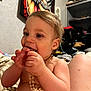 toddler, child, baby, necklace, shirtless, indoors, bed, blanket, furniture, wall_art, shelf, clutter, person, face, hand, curly_hair, happy, chewing, closeup, home