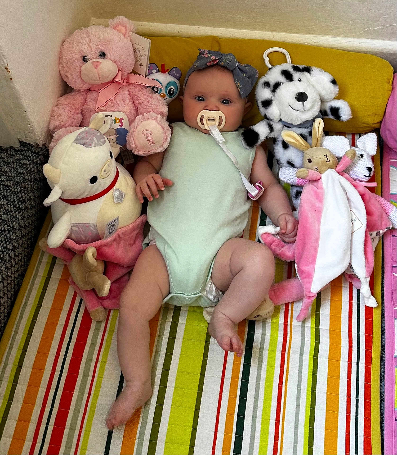 Millie is registered to the contest to win money with this photo: baby, infant, plush_toy, teddy_bear, stuffed_animal, pacifier, headband, striped_mat, colorful, child, cute, soft_toys, lying_down, indoors, pink, green_onesie, smiling, playtime, comfort, young