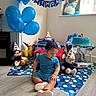 balloons, birthday, blue, cake, celebration, child, clothing, decorations, floor, indoor, mat, pacifier, party, party_hat, sitting, stuffed_animal, toddler, toy, toy_car, window