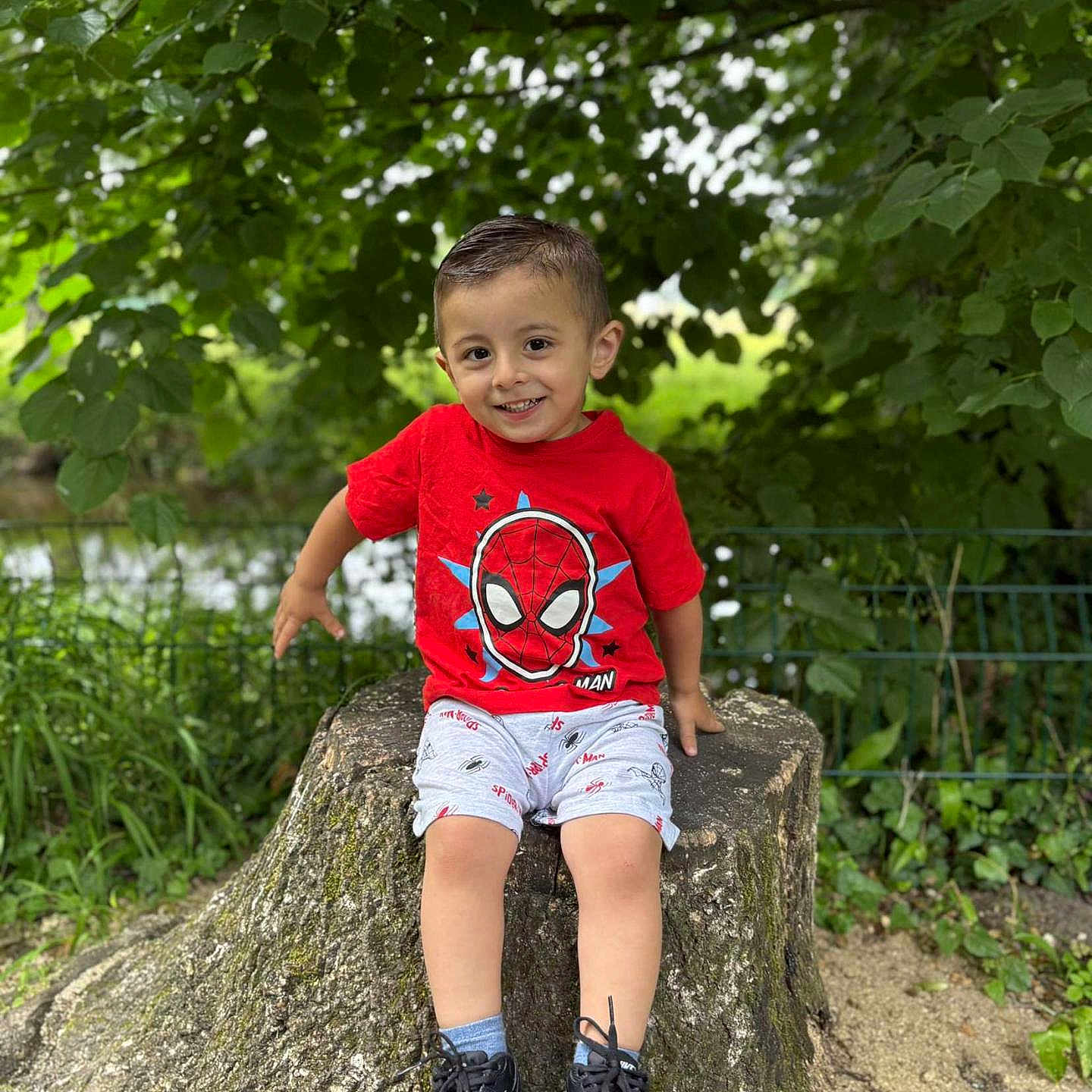 Akim a rejoint le concours — aidez-le/la à gagner de superbes lots ! boy, casual_clothing, child, cute, daylight, grass, greenery, happy, leaves, nature, outdoor, park, person, portrait, red_tshirt, shorts, smile, sneakers, spiderman, tree_stump