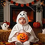 baby, ghost_costume, pumpkin_bucket, halloween, autumn_leaves, fireplace, jack_o_lantern, hay_bale, ghost_figurine, spider_web, costume, holiday_decor, indoors, child, festive, cute, smiling, floor, warm_lighting, seasonal