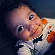 Sophia is registered to the contest to win money with this photo: baby, child, infant, portrait, smiling, happy, face, eyes, curly_hair, close_up, indoors, sitting, onesie, clothing, orange, pumpkin_pattern, soft_lighting, dark_background, photography, cute
