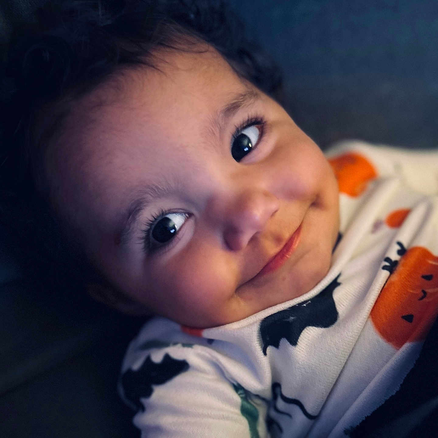Sophia is registered to the contest to win money with this photo: baby, child, infant, portrait, smiling, happy, face, eyes, curly_hair, close_up, indoors, sitting, onesie, clothing, orange, pumpkin_pattern, soft_lighting, dark_background, photography, cute