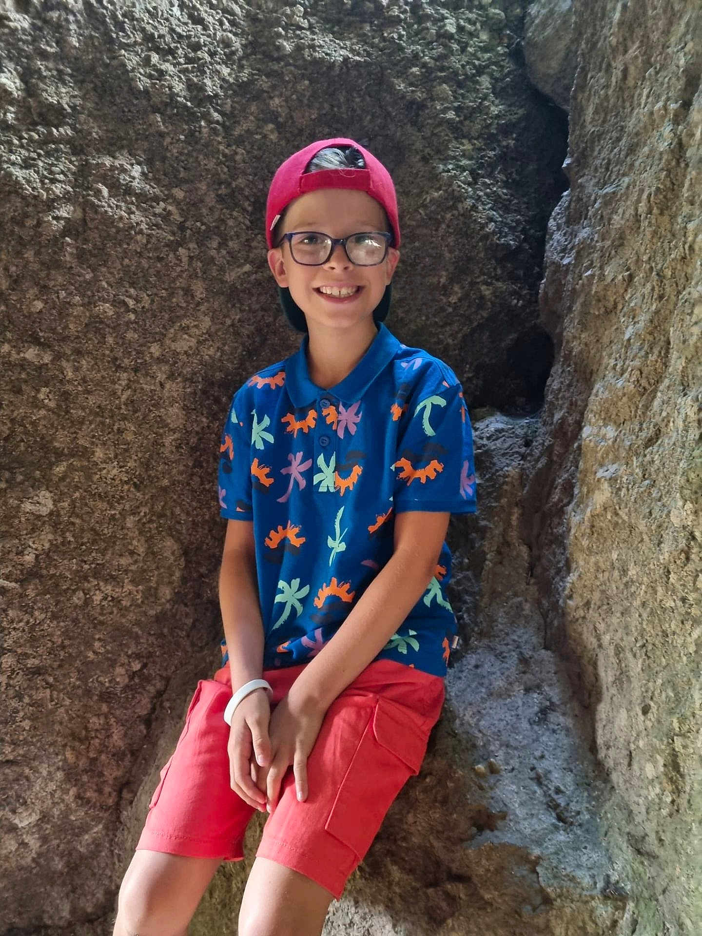 Kaïs participe au concours pour gagner de l'argent avec cette photo : child, boy, smile, glasses, red_cap, colorful_shirt, shorts, rock, cave, outdoor, happy, sitting, nature, casual_clothing, portrait, young, hands, light, shadow
