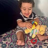 baby, child, toddler, smiling, sitting, stuffed_animal, toy, plush_toy, teething_ring, bed, bedspread, patterned_fabric, indoors, portrait, curly_hair, striped_clothing, infant, playtime, happy, feet