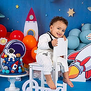 Kenzy participe au concours pour gagner de l'argent avec cette photo : toddler, child, smile, white_clothing, chair, birthday_cake, astronaut, rocket, balloons, space_theme, decorations, stars, planets, blue_background, orange_balloons, red_balloons, happy, celebration, first_birthday, party