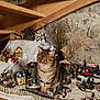 attic, brown_cat, cat, curious, decorations, fence, festive, figurines, holiday, indoor, miniature_village, ornaments, pine_cone, plants, sitting, stone_wall, tabby, toy_car, winter_scene, wooden_beams