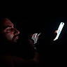 man, beard, face, person, cat, kitten, animal, pet, smartphone, phone, screen, darkness, night, low_light, portrait, profile, whiskers, fur, indoor, dramatic_lighting