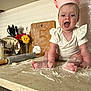 baby, child, chef_hat, kitchen, flour, rolling_pin, countertop, flowers, cutting_board, smiling, sitting, messy, cooking, baking, infant, white_clothing, happy, face, hands, toes