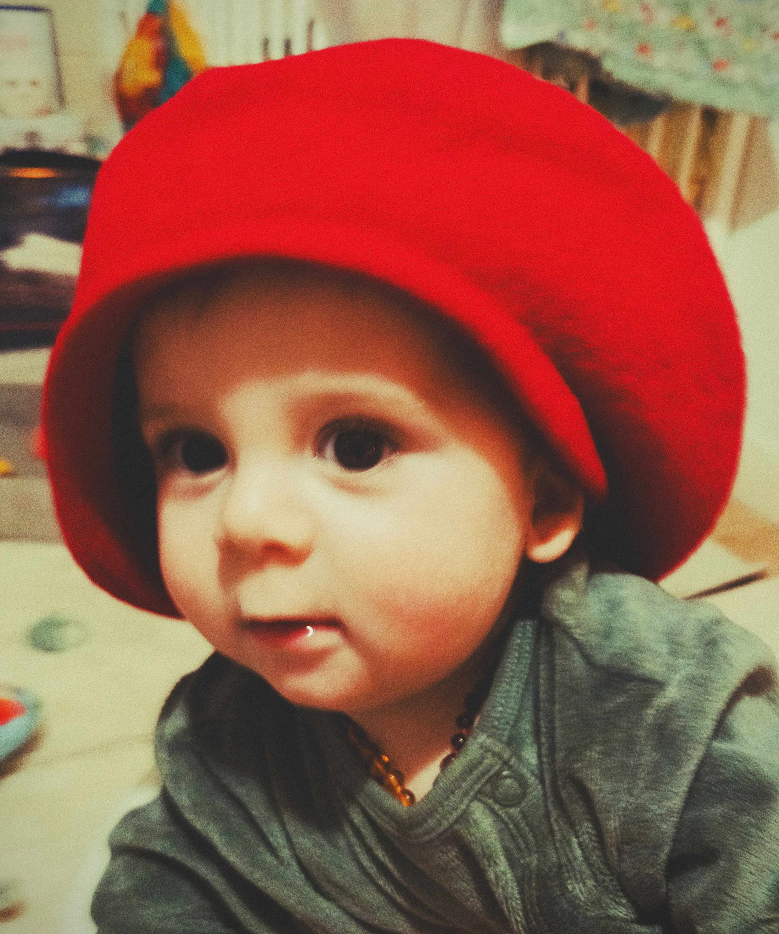 Marceau participe au concours pour gagner de l'argent avec cette photo : baby, child, red_hat, green_clothing, face, close_up, indoor, portrait, young_child, cute, curious, soft_focus, warm_colors, infant, expression, headwear, person, indoors, small_child, necklace