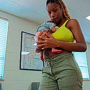 Mora joined the competition — help win amazing prizes! baby, infant, mother, parent, 抱_child, holding_baby, indoor, clinic, classroom, waiting_room, portrait, standing, braids, green_tank_top, cargo_pants, blanket, couch, chair, ceiling_tiles, window