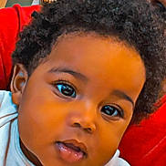 Zakai is registered to the contest to win money with this photo: baby, child, face, portrait, curly_hair, eyes, expression, infant, cute, young_child, red_background, white_clothing, skin, head, person, closeup, indoors, human, looking, curious