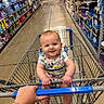baby, child, shopping_cart, supermarket, grocery_store, aisle, smiling, portrait, infant, floral_clothing, onesie, shopping, retail, store_shelves, household_products, paper_products, cart_handle, adult_hand, indoor, happy