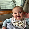 baby, child, infant, smiling, happy, portrait, car, car_seat, vehicle, indoors, travel, bib, heart_pattern, blue_clothing, pants, baby_carrier, seatbelt, window, cloudy_sky, face