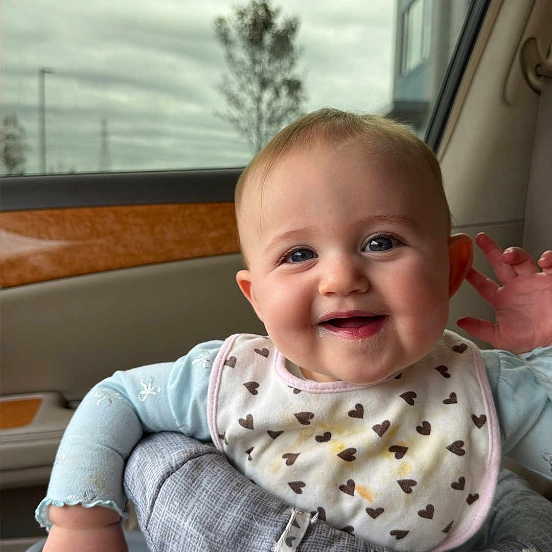 Siena is registered to the contest to win money with this photo: baby, child, infant, smiling, happy, portrait, car, car_seat, vehicle, indoors, travel, bib, heart_pattern, blue_clothing, pants, baby_carrier, seatbelt, window, cloudy_sky, face