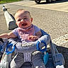 baby, child, infant, smiling, happy, outdoor, stroller, parked_car, pavement, sunlight, portrait, face, laughing, bib, purple_clothing, gray_pants, baby_toy, parking_lot, trees, summer