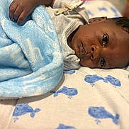 Shiloh is registered to the contest to win money with this photo: baby, infant, child, portrait, lying_down, blanket, blue_blanket, bed, bedding, face, eyes, looking_at_camera, dark_skin, black_hair, gray_clothing, cozy, indoors, close_up, sleeping_area, soft_lighting