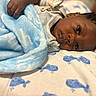 baby, infant, child, portrait, lying_down, blanket, blue_blanket, bed, bedding, face, eyes, looking_at_camera, dark_skin, black_hair, gray_clothing, cozy, indoors, close_up, sleeping_area, soft_lighting