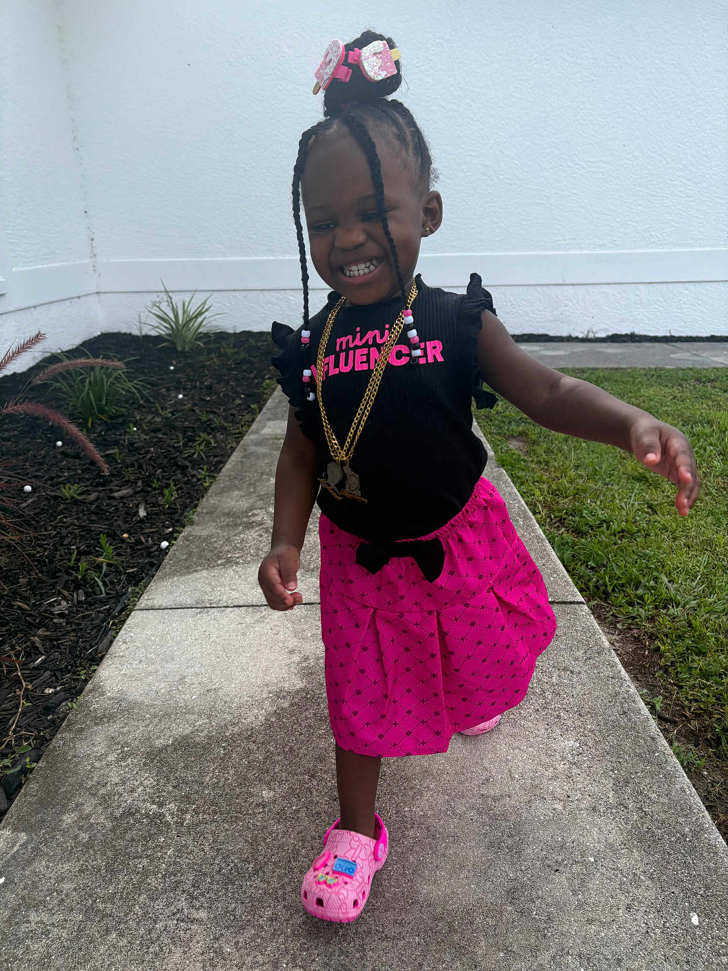 Miliana is registered to the contest to win money with this photo: child, girl, smiling, braids, pink_skirt, black_top, outdoor, sidewalk, grass, garden, happy, walking, footwear, accessories, necklace, hair_bun, casual_clothing, daylight, portrait, cute