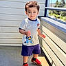 child, toddler, boy, standing, outdoor, balcony, sunlight, clothing, shorts, tshirt, red_shoes, cartoon, hair, smile, face, person, daylight, railing, concrete_floor, playful