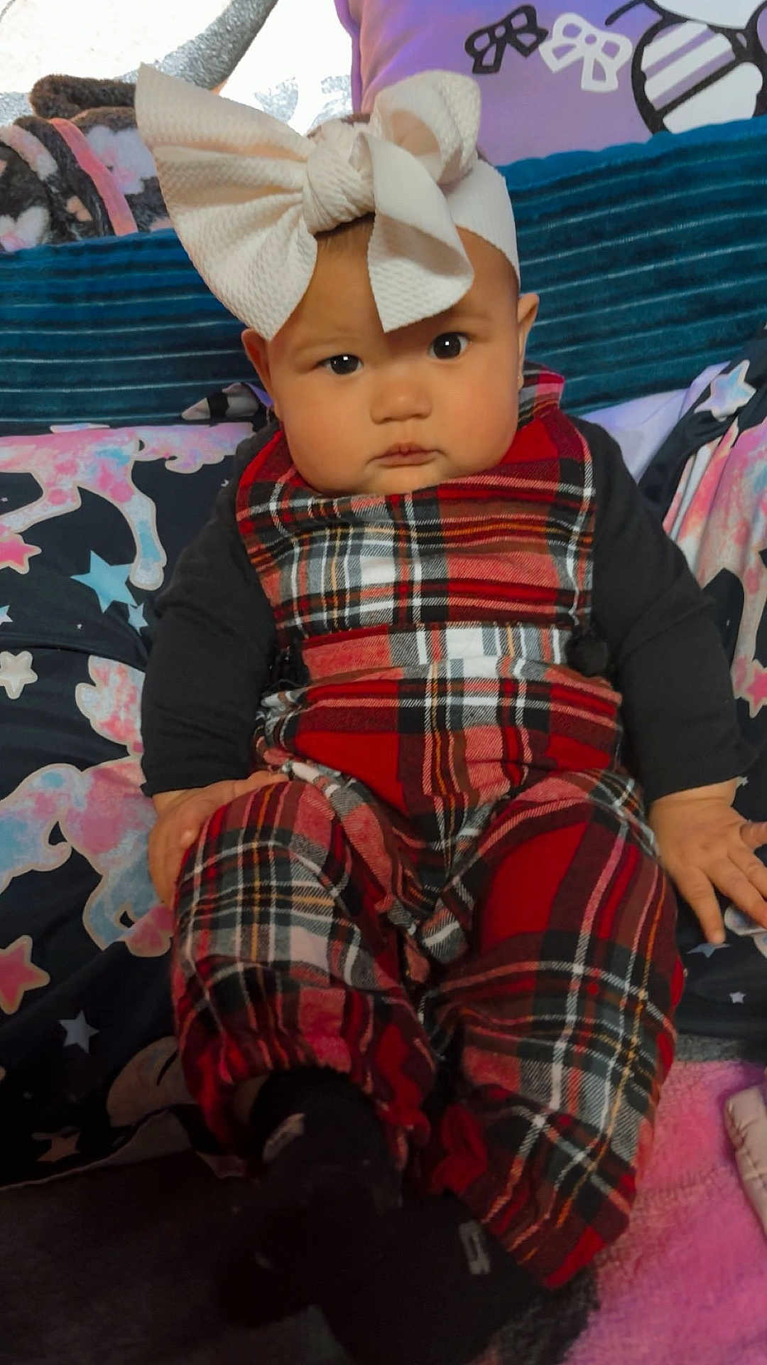 Kaylani joined the competition — help win amazing prizes! baby, child, headband, bow, plaid, overalls, clothing, sitting, cushion, blanket, indoor, face, person, cute, fashion, black_socks, expression, portrait, infant, colorful
