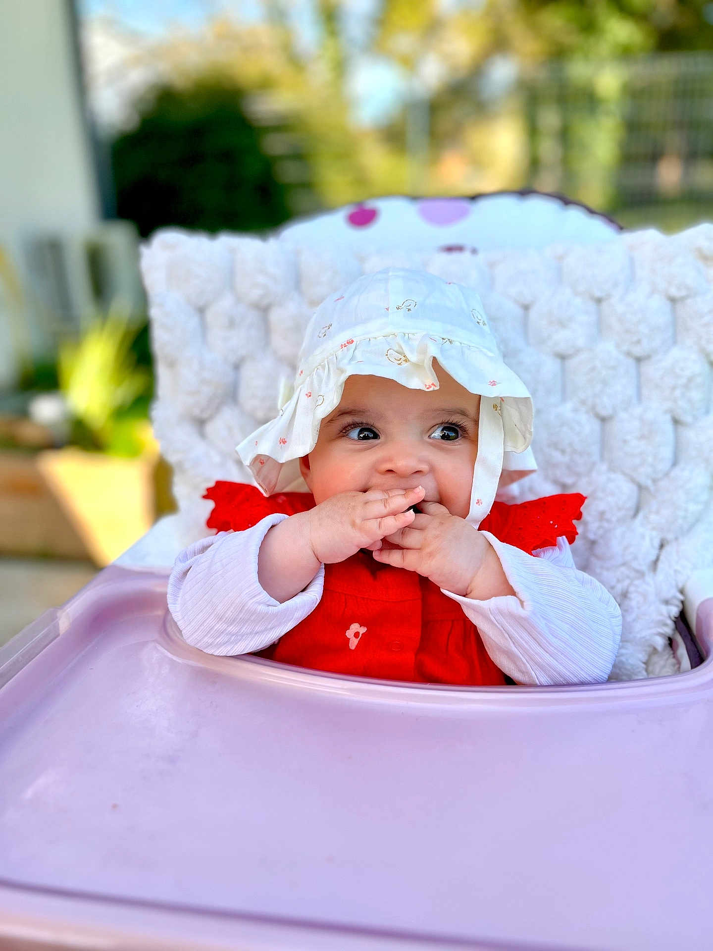 Esmée participe au concours pour gagner de l'argent avec cette photo : baby, child, high_chair, hat, red_clothing, white_hat, cute, infant, fingers_in_mouth, knitted_blanket, outdoor, daylight, portrait, happy, smiling, seat, young_child, face, person, baby_girl