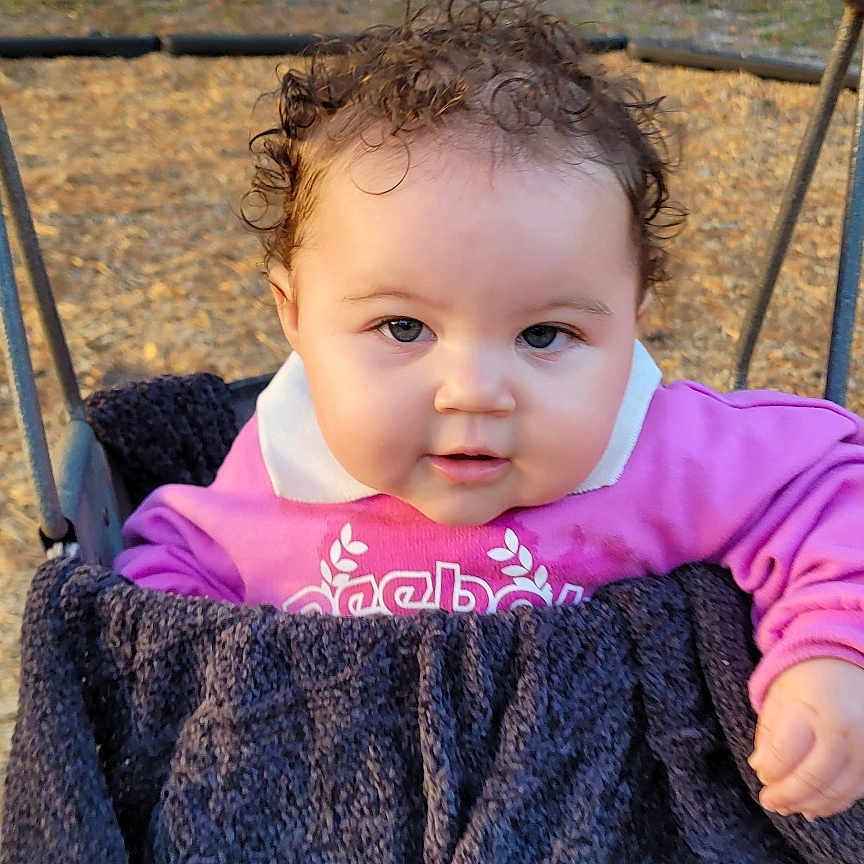 Maliah is registered to the contest to win money with this photo: baby, blanket, cheeks, child, cozy, curly_hair, eyes, face, infant, outdoors, park, photography, pink_sweater, playground, portrait, seat, swing, trees, white_fence, wood_chips
