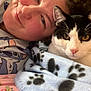 person, cat, black_and_white_cat, blanket, paw_prints, bed, pajamas, smiling, face, closeup, indoor, resting, cozy, cute, pet, animal, feline, human, comfort, relaxation