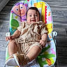 baby, child, infant, smiling, happy, portrait, seated, bouncer, activity_seat, knitted_outfit, beige_clothing, shoes, colorful_cushion, indoor, wood_floor, home, natural_light, face, playful, adorable