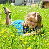 child, grass, yellow_flowers, blue_shirt, outdoor, smiling, happy, nature, sunlight, greenery, playful, face, person, casual_clothing, daylight, field, pigtails, shoe, meadow, portrait