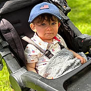 Carter joined the competition — help win amazing prizes! toddler, child, stroller, blue_cap, american_flag, blanket, outdoor, grass, shirt, patterned_clothing, seatbelt, baby, person, portrait, cute, sunlight, daylight, young_child, face, expression