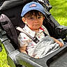 toddler, child, stroller, blue_cap, american_flag, blanket, outdoor, grass, shirt, patterned_clothing, seatbelt, baby, person, portrait, cute, sunlight, daylight, young_child, face, expression