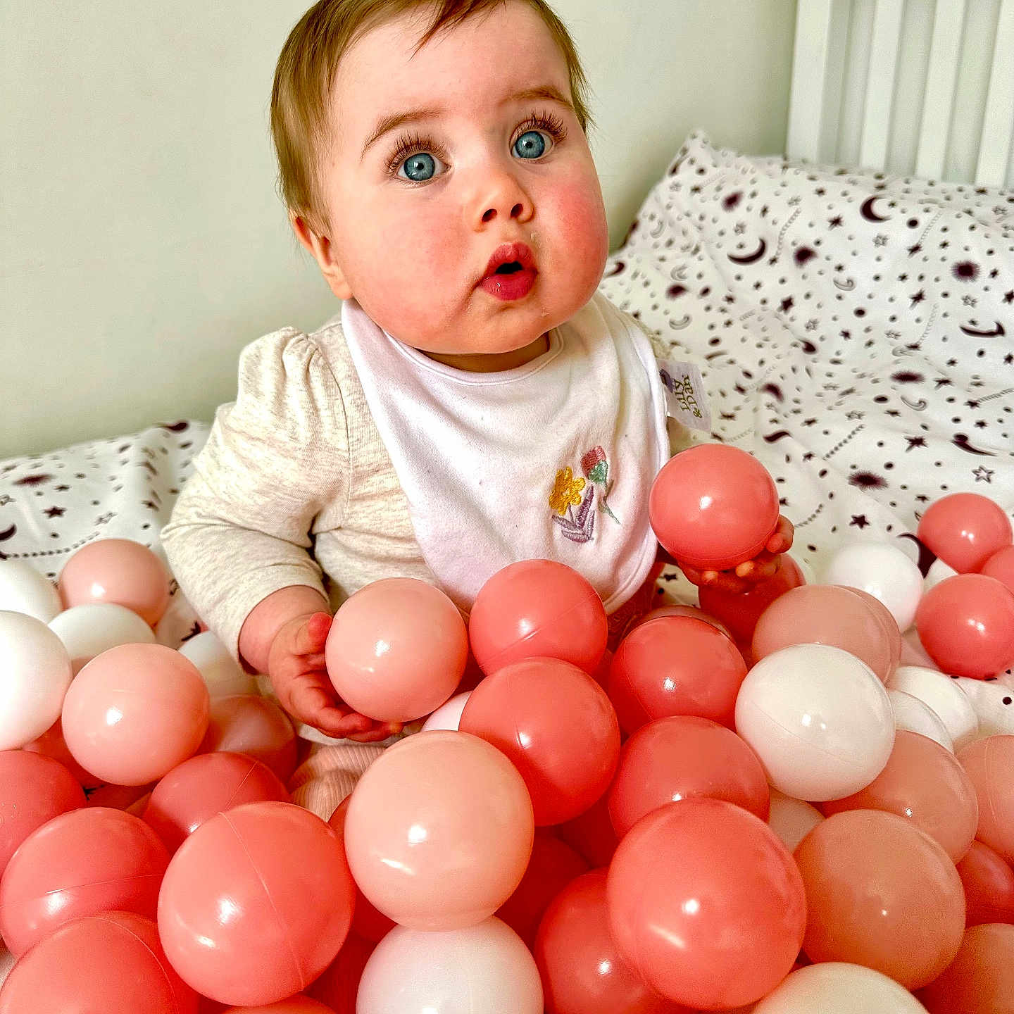 Aurora is registered to the contest to win money with this photo: baby, balls, bed, bib, blanket, blue_eyes, cheeks, child, clothing, cute, expression, face, indoor, patterned, person, pink, plastic_balls, sitting, toy, white