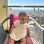 baby, balcony, building, chair, child, cityscape, cushion, daylight, dress, face, happy, infant, legs, outdoor, person, railing, sky, smile, sunlight, window
