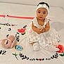 adorable, baby, child, curious, cute, decorative_blanket, floor, flower_design, headband, indoors, infant, looking_up, milestone_blanket, portrait, sitting, small_object, toy, white_dress, white_socks, young_child