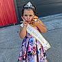 child, girl, floral_dress, tiara, sash, plush_toy, stuffed_animal, peace_sign, pout, portrait, outdoor, concrete, pavement, red_wall, metal_shutter, white_shoes, bow_shoes, pageant, crown, celebration