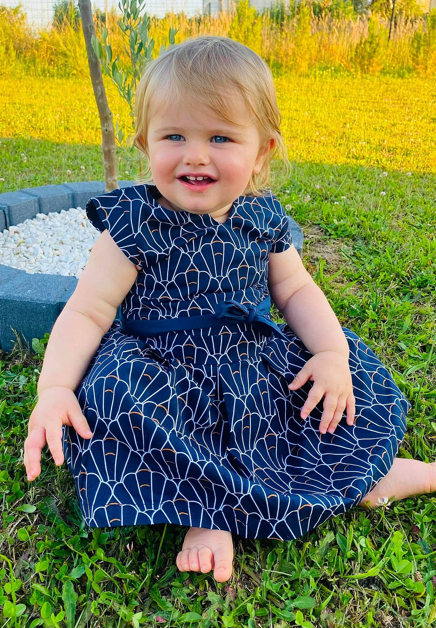 Taïs participe au concours pour gagner de l'argent avec cette photo : baby_toddler_clothing, dress, facial_expression, grass, green, hairstyle, happy, joy, leaf, nature, one_piece_garment, people_in_nature, person, plant, product, skin, smile, standing, sunlight, toddler