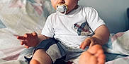 Lino participe au concours pour gagner de l'argent avec cette photo : baby, child, toddler, pacifier, barefoot, bed, blanket, white_shirt, shorts, indoor, curly_hair, face, expression, sitting, foot, hand, clothing, young, portrait, light