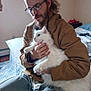 Snowball is registered to the contest to win money with this photo: beard, bed, bedroom, blue_quilt, brown_jacket, calm, casual_clothing, cat, furry, glasses, hands, holding, indoor, man, pet, quilt, seated, sweatpants, white_cat, window