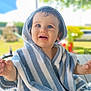 baby, blue_and_white, blurred_background, child, curious_expression, daylight, drool, face, greenery, hooded_towel, outdoor, person, playground, portrait, striped_towel, summer, sunlight, toddler, wooden_table, young_child